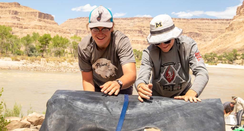 Two people roll up a large tarp. Behind them is a river and very tall red canyon walls